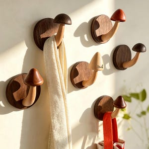 Könnte beinhalten: Eine Ansammlung von sechs pilzförmigen Wandhaken aus Holz. Jeder Haken besteht aus einer dunklen, runden Basis und einem helleren Holzstiel mit einem Hut. Ein Pilzhut ist mit dem Namen Noah graviert. Ein weißer Schal und ein roter Riemen hängen daran.