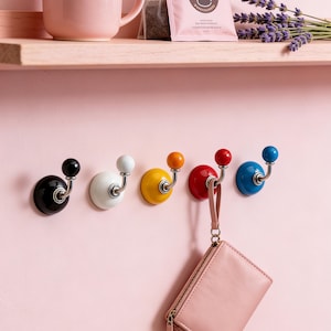 Könnte beinhalten: Sechs farbenfrohe Wandhaken mit rundem Knaufdesign. Die Haken sind schwarz, weiß, gelb, rot und blau. Jeder Haken hat einen silberfarbenen gebogenen Arm und ist an einer rosa Wand befestigt. Eine rosa Handtasche hängt an einem der Haken.