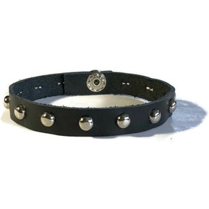 May include: Black leather bracelet with silver-colored studs. The bracelet has a snap closure and is adorned with multiple round, silver-toned studs. The leather appears to be smooth and the studs are evenly spaced around the band. A stylish accessory.