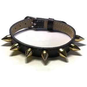 May include: Black leather bracelet with gold-colored spikes. The bracelet has a silver-colored buckle and is labeled "GENUINE LEATHER" on the inside. The spikes are evenly spaced around the bracelet.