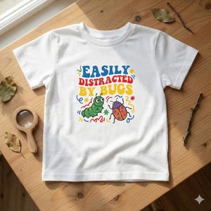 May include: White t-shirt with the text "EASILY DISTRACTED BY BUGS" in blue, red, and yellow. The shirt features a green caterpillar and a colourful ladybug graphic. The shirt is displayed on a wooden surface.