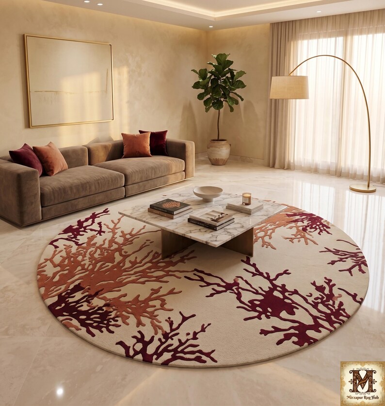 Puede incluir: Una alfombra circular grande con una base de color crema y un intrincado patr&oacute;n de ramas que recuerda al coral en tonos burdeos intenso y naranja quemado. La alfombra est&aacute; colocada sobre un suelo pulido bajo una mesa de centro de m&aacute;rmol.