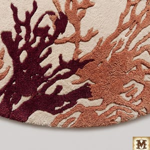 Puede incluir: Una alfombra circular con una base de color crema y un dise&ntilde;o copetudo en relieve de estructuras ramificadas similares al coral en tonos burdeos intenso y naranja terracota apagado. La textura es densa y afelpada, creando contraste.