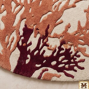 Puede incluir: Una alfombra circular con textura de pelo largo y mechones sobre una base de color crema. El dise&ntilde;o muestra un patr&oacute;n intrincado de ramas que recuerdan al coral en tonos naranja quemado y granate profundo, creando un fuerte contraste visual.