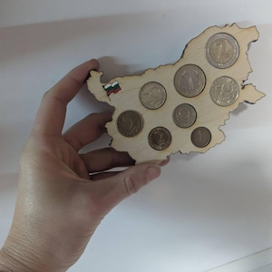 May include: A wooden coin holder in the shape of Bulgaria, with coins of various denominations placed in the cutouts. The holder has a small Bulgarian flag and is held by a hand against a white background.