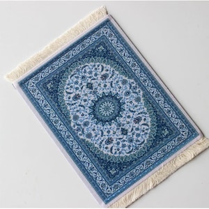 Puede incluir: Una pequeña alfombra rectangular con un detallado estampado floral en tonos azules y blancos. La alfombra presenta un diseño de medallón central y un borde con intrincados adornos. Flecos color crema bordean la parte superior e inferior.