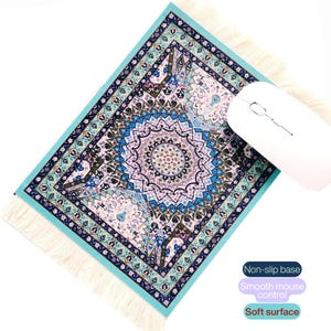 Pode incluir: Tapete de rato retangular decorativo com um design detalhado e colorido de tapete oriental. O tapete de rato apresenta um padrão de medalhão central em tons de azul, rosa e verde, com uma borda turquesa e bordas com franjas. Um rato sem fios branco está em cima.