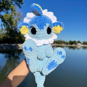 May include: A light blue, soft plush toy featuring a creature with large black eyes, a white ruffled collar, and yellow-tipped ears. The body has darker blue spots on the limbs and a fish-like tail, all crafted from a textured, fuzzy yarn material.