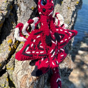 May include: A plush dragon toy crafted from red, black, and white yarn. The creature features a long neck, a prominent blue eye, and textured wings with black and white accents. The body is constructed with soft, fuzzy yarn in a vibrant red hue.