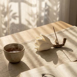 May include: A tranquil still life composition showing a white ceramic flower incense holder with a lit incense stick, a small ceramic cup, and an open book with spectacles. The items are arranged on a beige table runner, illuminated by soft sunlight.