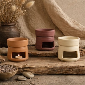 Può includere: Tre bruciatori di olio in ceramica nei toni del beige, bordeaux e crema sono su una superficie di legno. Il bruciatore beige ha un lumino acceso all'interno. Sono presenti anche una piccola ciotola di lavanda e pietre decorative.