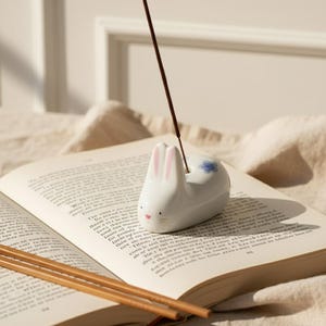 May include: A white ceramic bunny incense holder with pink ears and a blue floral accent. The incense holder is on an open book with several incense sticks. The bunny has a small pink nose and is resting on the book.