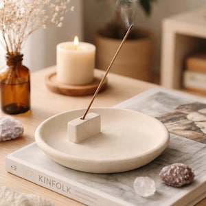 Op de afbeelding: Een beige keramische wierookhouder met een brandende wierookstok, die een wolkje rook afgeeft. Een brandende kaars en een decoratieve vaas met gedroogde bloemen staan op de achtergrond. De scène speelt zich af op een houten tafel met een boek.