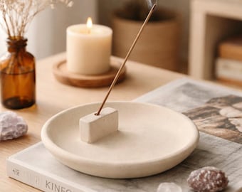 May include: A beige ceramic incense holder with a lit incense stick, emitting a wisp of smoke. A lit candle and a decorative vase with dried flowers are in the background. The scene is set on a wooden table with a book.