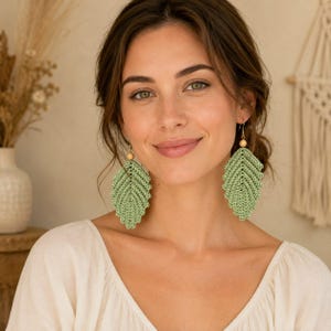 May include: A pair of sage green crochet earrings shaped like leaves. Each earring features a textured, pointed leaf design crafted from fine thread, accented with a small, round, light-colored wooden bead at the top where the metal hook attaches.