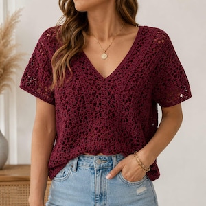 May include: A deep burgundy short-sleeved top featuring an intricate open-knit crochet pattern with a V-neckline. The garment is paired with light blue denim jeans, a gold pendant necklace, and a gold chain bracelet on the wrist.