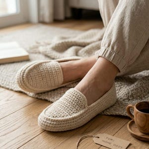 以下が含まれることがあります： クリーム色の、テクスチャードデザインのクロシェ編みスリッパ。 スリッパはローファーのスタイルで、ニュートラルカラーのパンツと組み合わせています。 木製のコーヒーカップが木製のコースターの上にあり、タグには「FOR: HOME COZY」と書かれています。