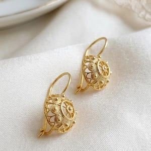 Puede incluir: Un par de pendientes de oro con un diseño circular abombado de estilo filigrana, con intrincados patrones de alambre, pequeñas esferas en relieve y un motivo central en forma de rueda, sujetos mediante ganchos curvos.