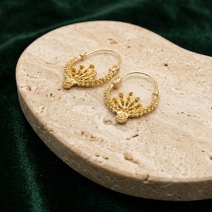 May include: A pair of gold-coloured hoop earrings. Each earring has a detailed, textured design with a fan-like shape at the bottom. The earrings are displayed on a light-coloured, textured surface against a dark green background.