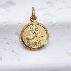 May include: A round gold pendant featuring a raised relief design of a crowned figure holding a child. The pendant has a smooth, polished circular border and a small integrated bail at the top for attachment to a chain or necklace.
