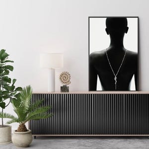 May include: A framed monochrome photograph of a person's back, adorned with a silver necklace. The necklace features a pendant. The photograph hangs above a black cabinet with vertical detailing. A white lamp and decorative items are on the cabinet.