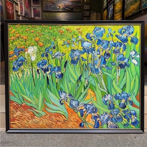 以下が含まれることがあります： 鮮やかな庭の風景を描いた額装された油絵の複製画。濃い青色のアイリスが多数と、白いアイリスが1輪描かれています。長い緑の葉、背景のオレンジ色の花、そして質感のあるオレンジ色の土が特徴的な構図です。