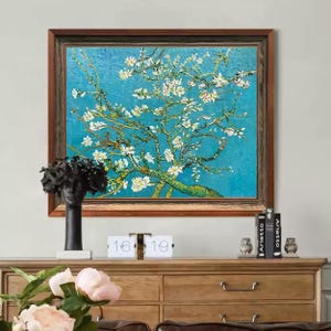 以下が含まれることがあります： 青い背景に咲き誇るアーモンドの木の額入り絵画。白い花と茶色の枝が特徴で、濃い茶色の木製フレームに収められています。黒い彫刻と装飾品が木製のキャビネットの上に置かれています。