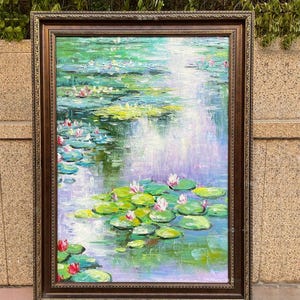 以下が含まれることがあります： 青、紫、白の水面に鮮やかな緑のスイレンの葉とピンクの花が浮かぶスイレンの池を描いたキャンバス地の油絵で、装飾が施されたダークブラウンの長方形の木製フレームに収められています。