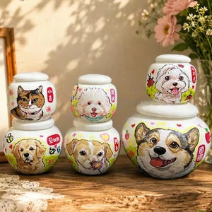 以下が含まれることがあります： 蓋付きの白い陶器製ジャー5個セット。三毛猫、ゴールデンレトリバー、コーギーなど、様々な犬や猫の品種が手描きで描かれており、小さなハートや花のモチーフが添えられています。