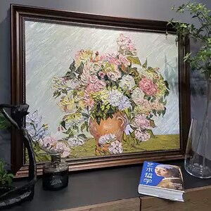 以下が含まれることがあります： ピンク、黄色、白の花が活けられた花瓶の油絵。絵画はテクスチャ加工が施され、淡い緑色の背景に配置されています。日本語のテキストが入った小さな本とガラス瓶が絵の前にあります。