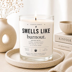 May include: A lit scented candle in a clear glass jar with the text "SMELLS LIKE burnout." The candle sits on a speckled ceramic plate. The text includes "INFUSED WITH: Coffee, unread emails and saying 'I'm fine' on 3 hours sleep."
