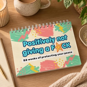 May include: A cream-coloured spiral-bound notebook with the text "Positively not giving a F*ck" in blue and white. The notebook features pink, green, and yellow abstract designs. The text "52 weeks of protecting your peace" is also visible.