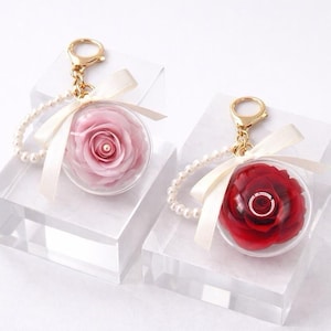 May include: Two keychains, one with a pink rose and the other with a red rose, encased in clear spheres. Each keychain has a gold clasp, a pearl bead chain, and a cream-colored ribbon. The roses are displayed on clear acrylic blocks.