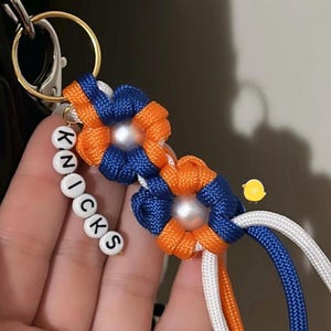 May include: A keychain featuring two flower-shaped charms woven from blue and orange paracord with a central white pearl-like bead. A string of white alphabet beads spells out KNICKS, attached to a gold-toned metal ring and a lobster clasp.