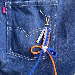 May include: A keychain featuring a braided cord in blue, white, and orange, attached to a gold-tone lobster clasp and a silver safety pin. A short strand of white beads with the black text KNICKS hangs from the clasp, and the cord ends in a bow.