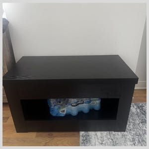 May include: A black wooden storage bench with a rectangular shape. The bench has a lower shelf that holds several bottles of water. The bench is placed on a wooden floor and a patterned rug.