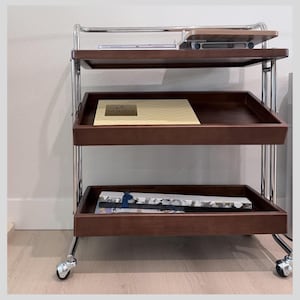 May include: A three-tiered serving cart with a chrome frame and dark brown wooden trays. The top tray holds a wooden cutting board and a laptop. The middle tray has a box with the word "GODIVA" on it. The bottom tray has a box with a blue ribbon.