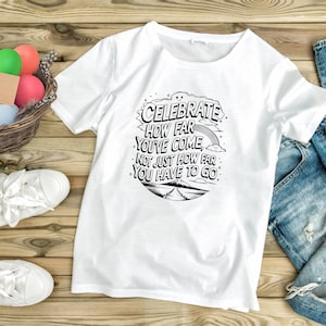 May include: White t-shirt with a black graphic that reads "Celebrate How Far You've Come, Not Just How Far You Have to Go." The design includes a rainbow, clouds, and a mountain scene. The shirt is on a wooden surface.