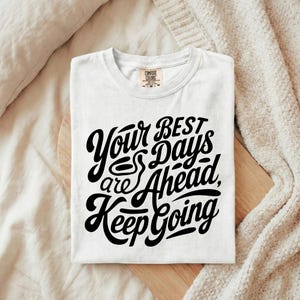 May include: White t-shirt featuring the black text "Your Best Days are Ahead, Keep Going". The shirt is folded on a wooden board. A neutral-toned blanket is in the background.