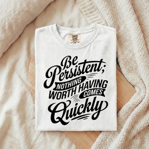 Puede incluir: Camiseta blanca con el texto negro "Be Persistent; Nothing Worth Having Comes Quickly". La camiseta doblada está colocada sobre una tabla de madera. Una manta de color crema está en el fondo.