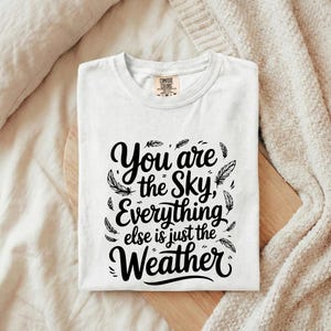 May include: A white short-sleeved t-shirt featuring black text that reads You are the Sky, Everything else is just the Weather. The design is surrounded by small black feather illustrations. The shirt has a Comfort Colors brand label at the neck.