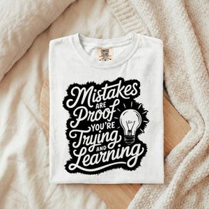 May include: White t-shirt featuring the quote "Mistakes are Proof You're Trying and Learning" in black script, with a lightbulb graphic. The shirt is folded on a wooden board.