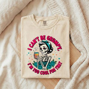 Może przedstawiać: Beżowy t-shirt z okrągłym dekoltem i grafiką szkieletu w turkusowej opasce, trzymającego szklankę z pomarańczowym płynem. Napis głosi I CAN'T BE GRUMPY, I'M TOO COOL FOR THAT. Wzór zawiera turkusowe róże oraz kolorowe gwiazdki.