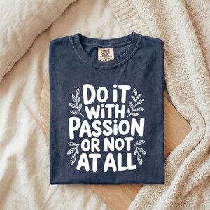 May include: A navy blue t-shirt with the white text "Do it with passion or not at all". The text is framed by leaf designs. The t-shirt is folded on a wooden board.