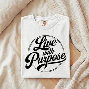 May include: White t-shirt with the words "Live with Purpose" in a black, circular design. The shirt is folded neatly on a wooden surface, with a cream-colored blanket in the background. The shirt is made by Comfort Colors.