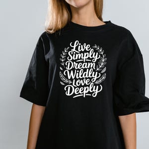 以下が含まれることがあります： 白い文字で「Live Simply, Dream Wildly, Love Deeply」と書かれた黒いTシャツ。葉のリースで囲まれています。テキストは筆記体です。Tシャツは半袖です。