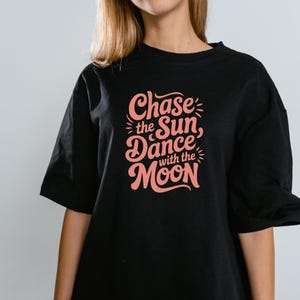 Puede incluir: Camiseta negra con el texto "Chase the Sun, Dance with the Moon" en coral, con una fuente de estilo retro. La camiseta de manga corta tiene cuello redondo. El diseño presenta gráficos estilizados del sol y la luna.