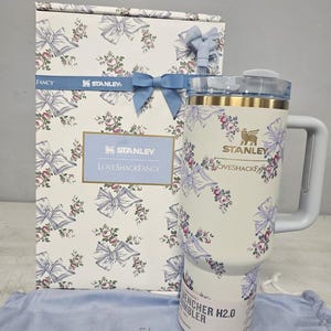 May include: A white Stanley Quencher H2.0 tumbler with a floral and bow design, matching a gift box. The tumbler has a handle and a lid. The box and tumbler feature the text "Stanley" and "LoveShackFancy".