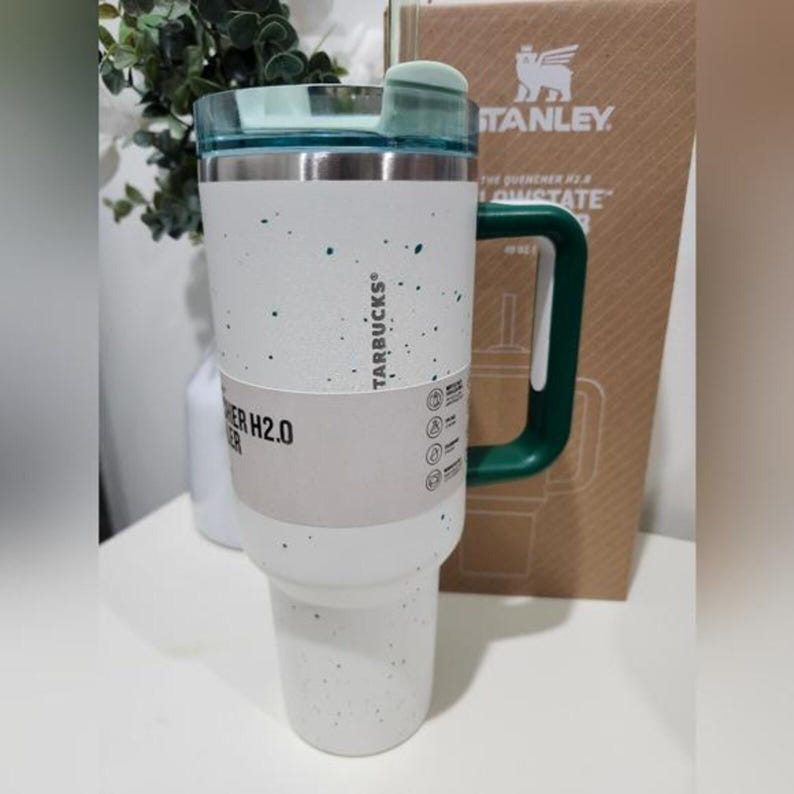 May include: A white and speckled Starbucks tumbler with a green handle and lid. The tumbler has a stainless steel rim and a white band with text. The tumbler is approximately 1.13 liters.