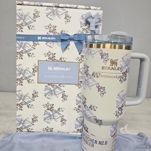 May include: A white Stanley Quencher H2.0 tumbler with a floral and bow design, matching a gift box. The tumbler has a handle and a lid. The box and tumbler feature the text "Stanley" and "LoveShackFancy".